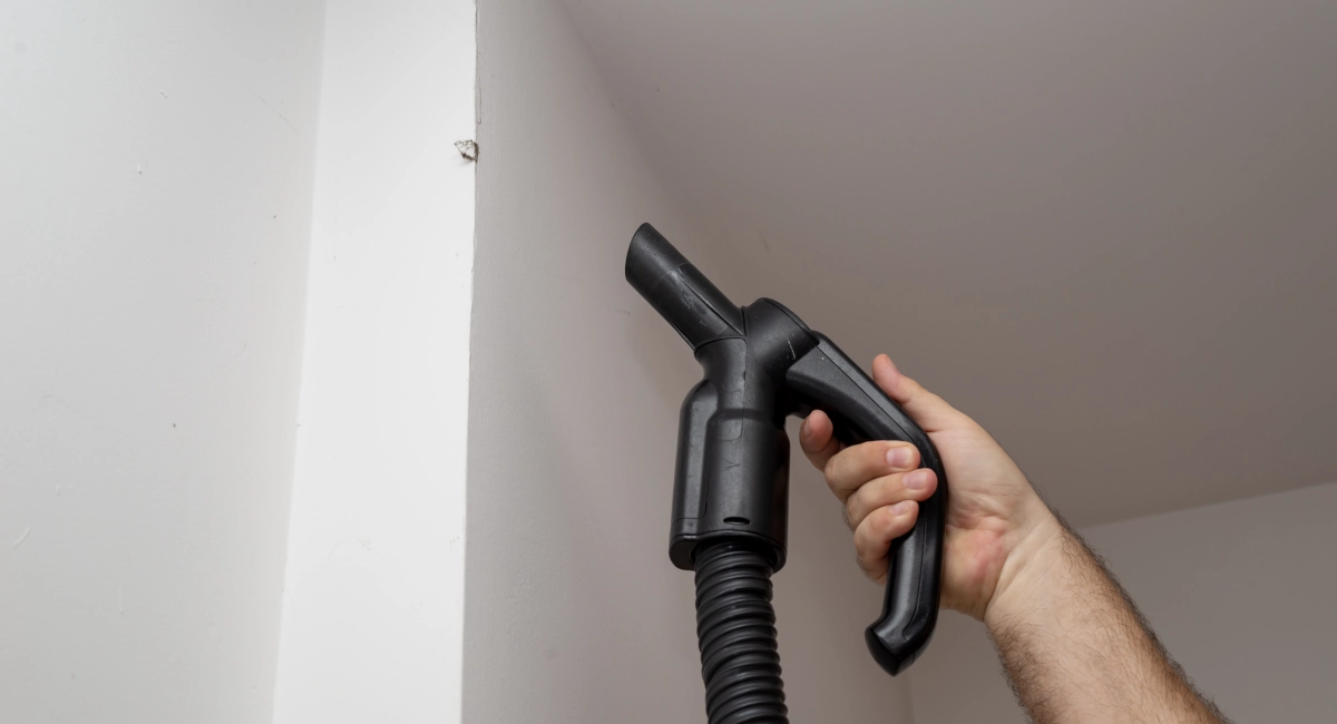 A person holds up a vacuum nozzle to a bit of debris on the wall