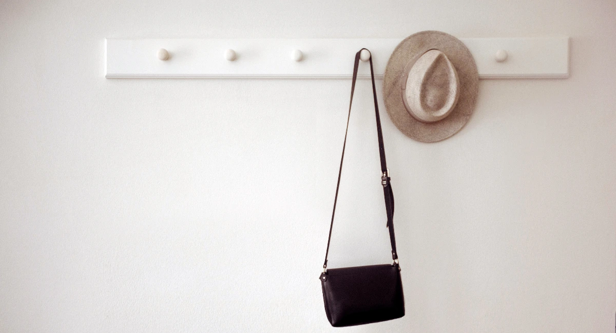 Stylish hat and small black purse hanging on white pegs on wall in cozy room
