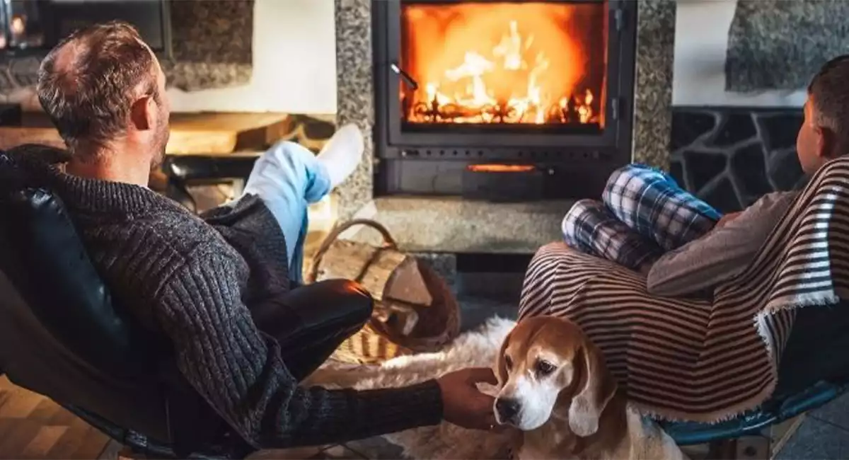 Man, woman, and their dog relaxing in recliners in front of a lit fireplace.