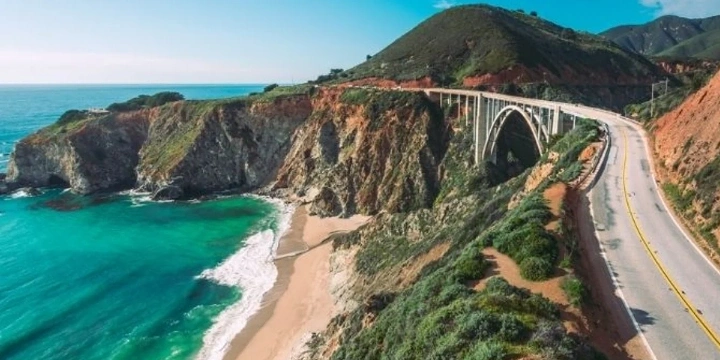 View of the Pacific Coast Highway showing blue water and sand next to it with a bridge ahead